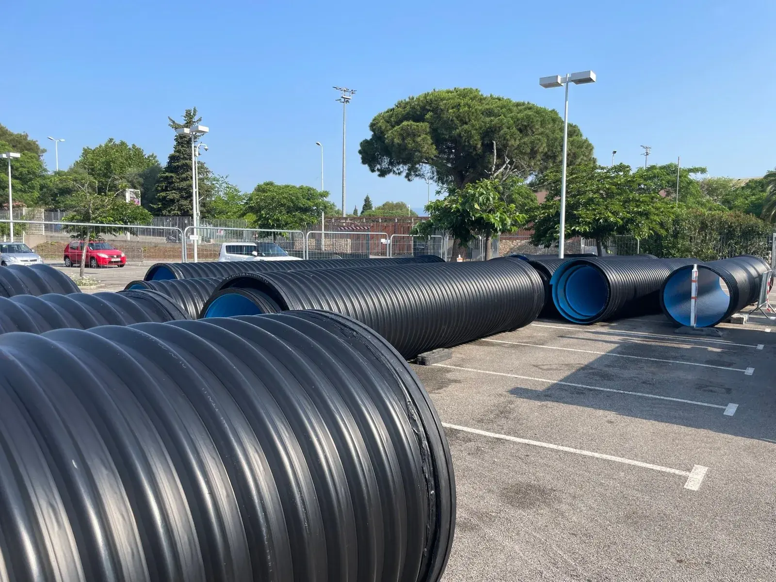 Black reinforced-profile HDPE pipes with blue interior, lined up in a car park before installation